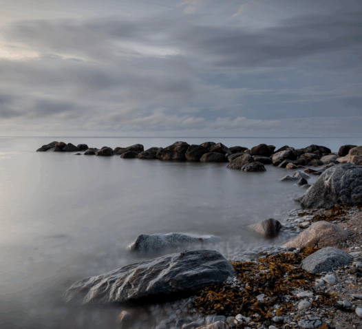 Stenar vid en kust med molnig himmel och lugnt vatten.