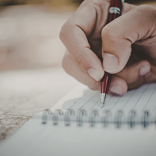 A hand holds a red pen, writing on a spiral-bound notebook with lined paper.